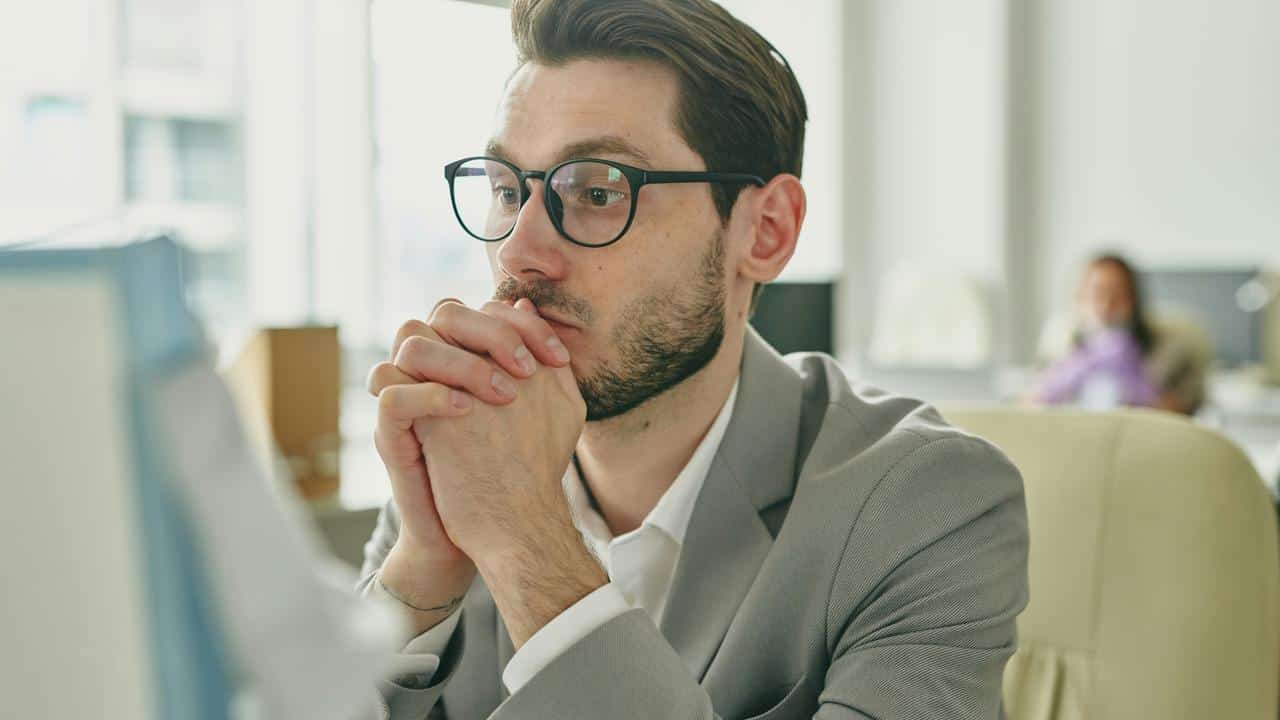 A pensive man in glasses resting his chin on clasped hands at a computer.