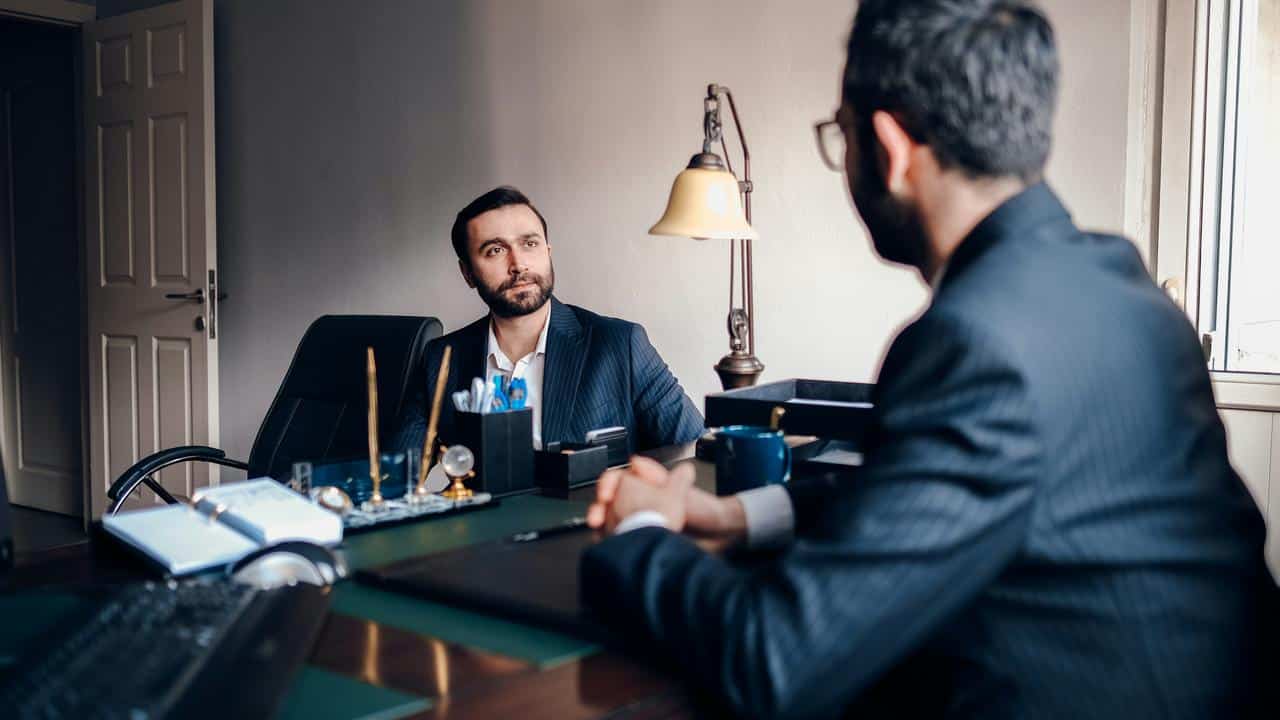 A man in a suit sitting across from another at a desk.