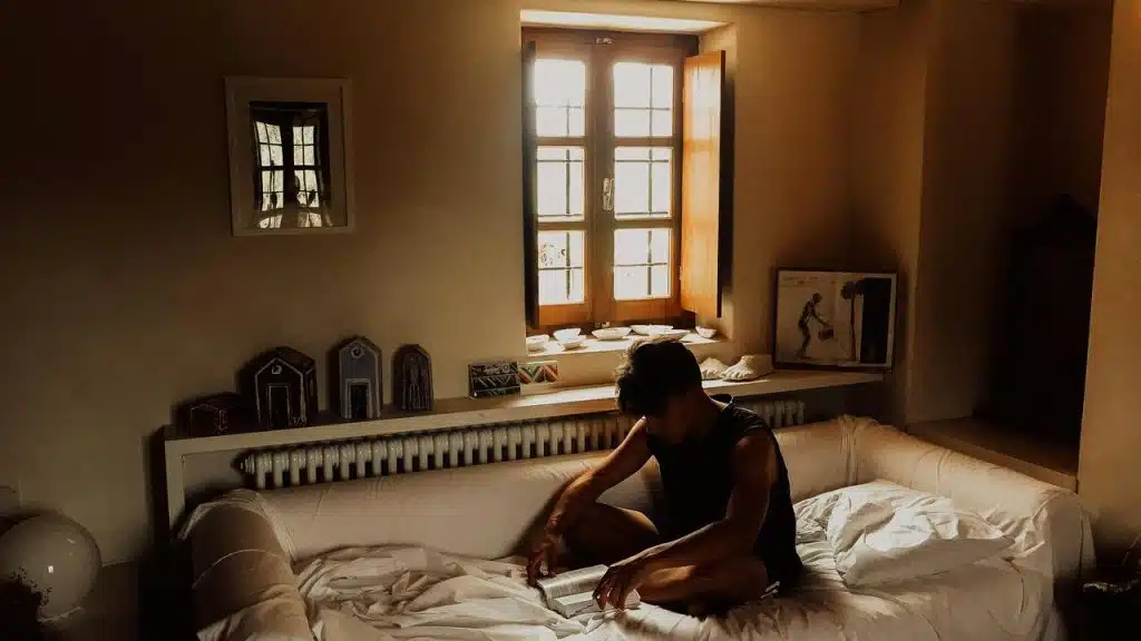 A man reading cross‑legged on a white sofa beneath a sunlit window.
