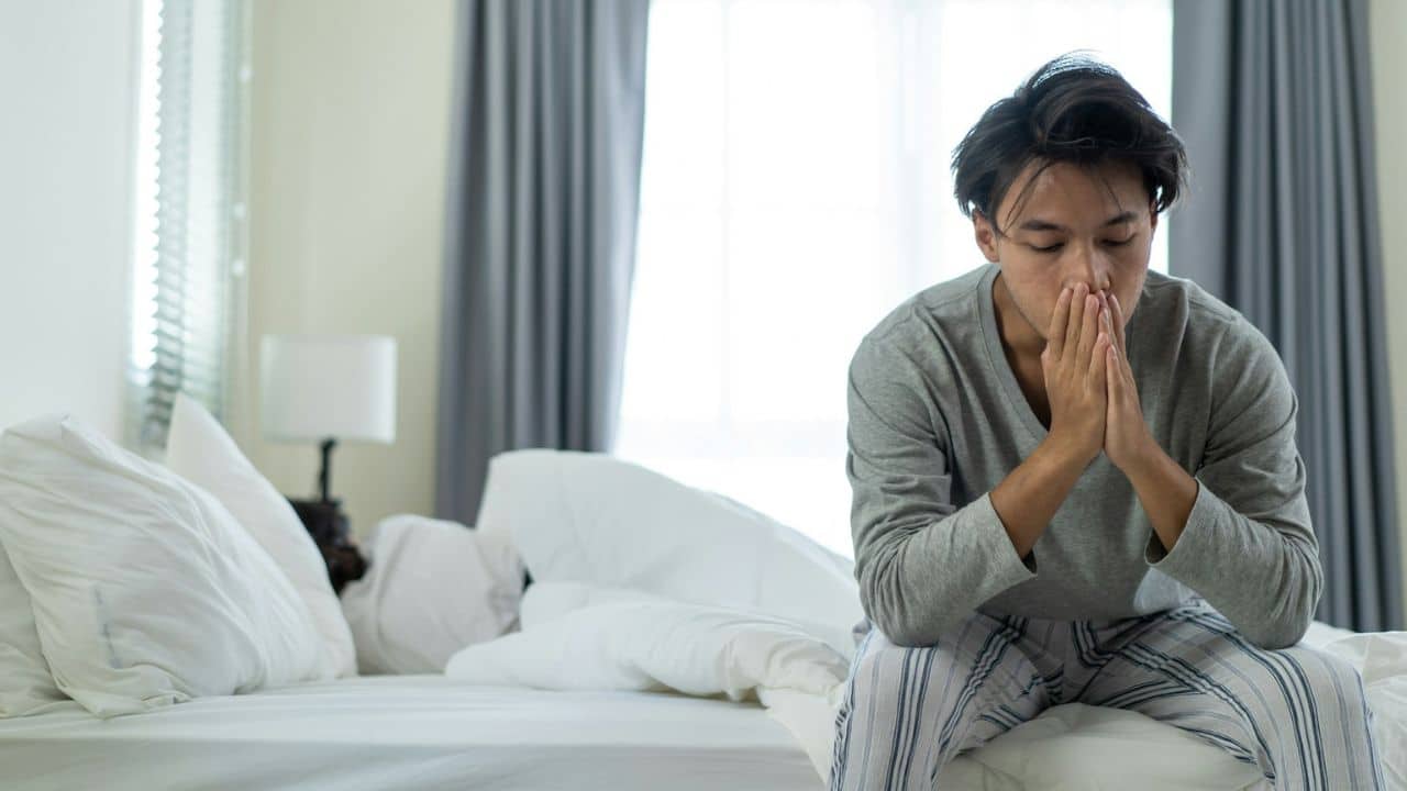 A man sits on a bed with his hands clasped, looking down, appearing distressed.
