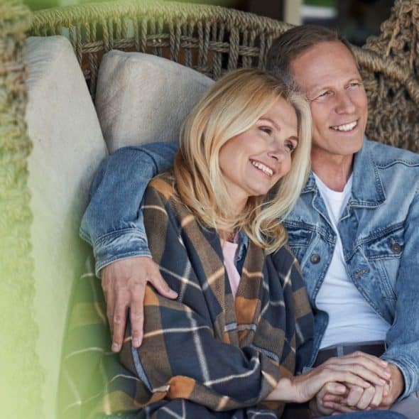 A smiling blonde woman rests her head on a man's shoulder, holding his hand, both seated outdoors.