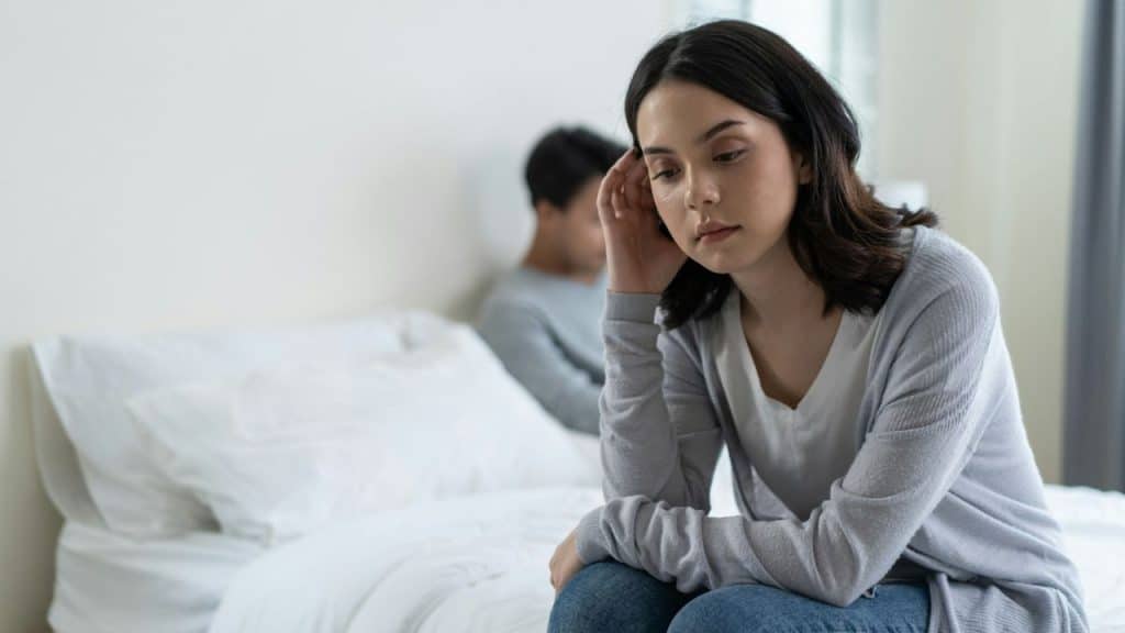 A thoughtful woman sits on a bed, looking down, with a man in the background.