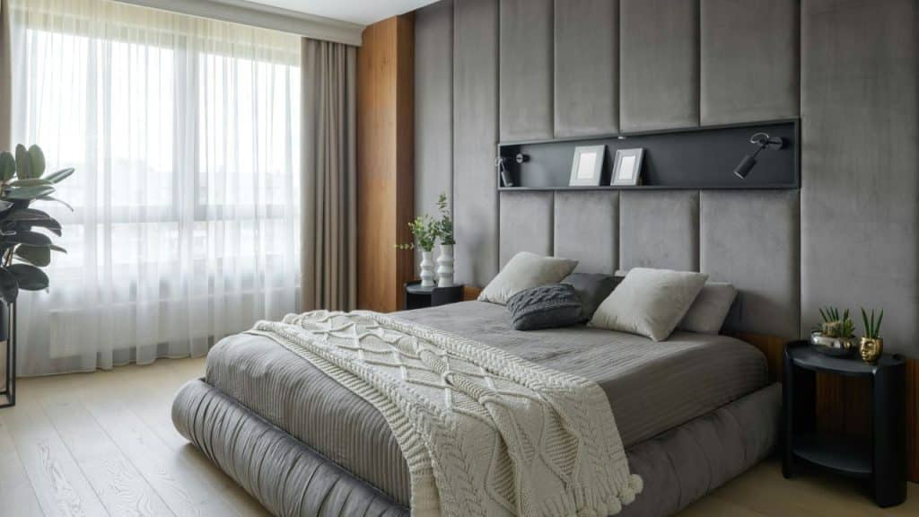 A modern bedroom with a large bed, gray padded headboard, and a window with sheer curtains.