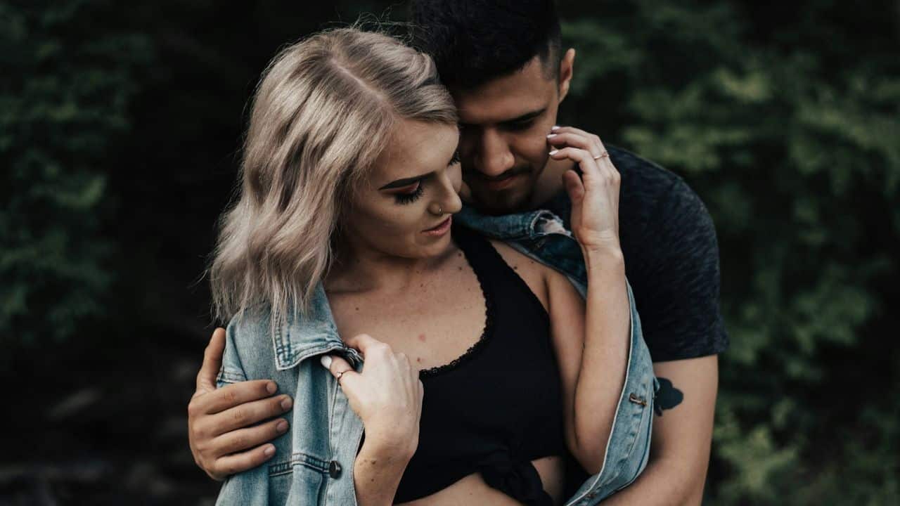 A man embraces a woman from behind, both looking down with a gentle expression.