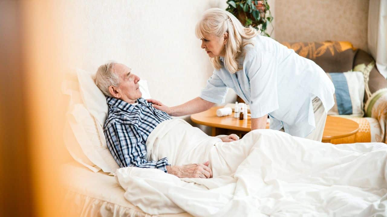 A woman in a light blue shirt leans over a man lying in bed, touching his shoulder.
