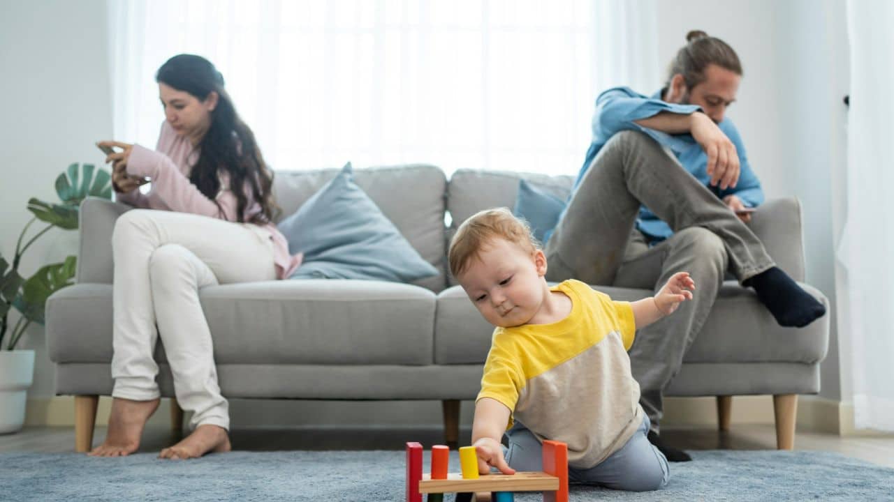 A baby plays on the floor while parents ignore them, engrossed in phones on the sofa.