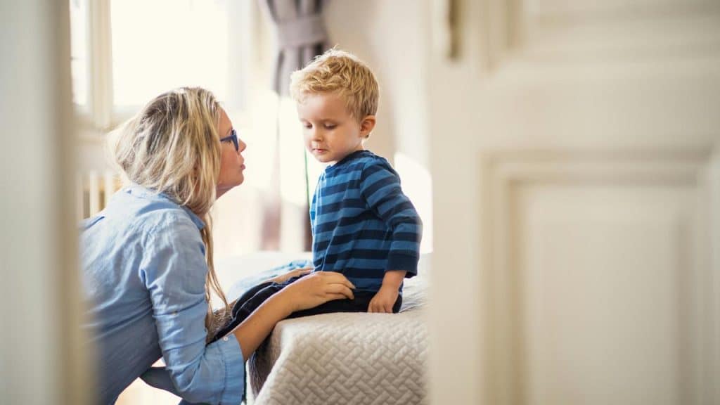 A mother kneeling beside her young son on a bed.