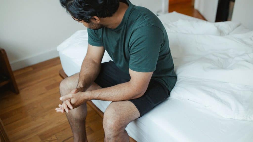 A man in a green shirt and black shorts sits on a bed, looking down.