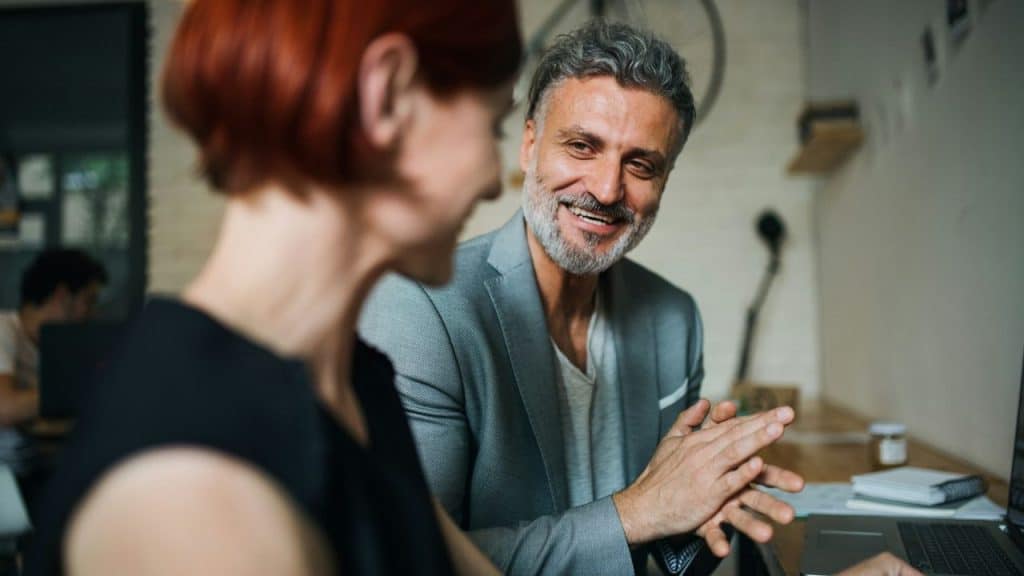 A man with gray hair and a beard smiles at a woman with red hair, at a desk.