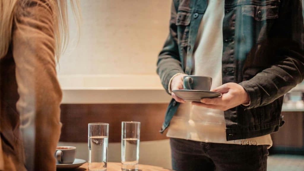 A person holding a coffee cup and saucer beside a table with glasses of water.