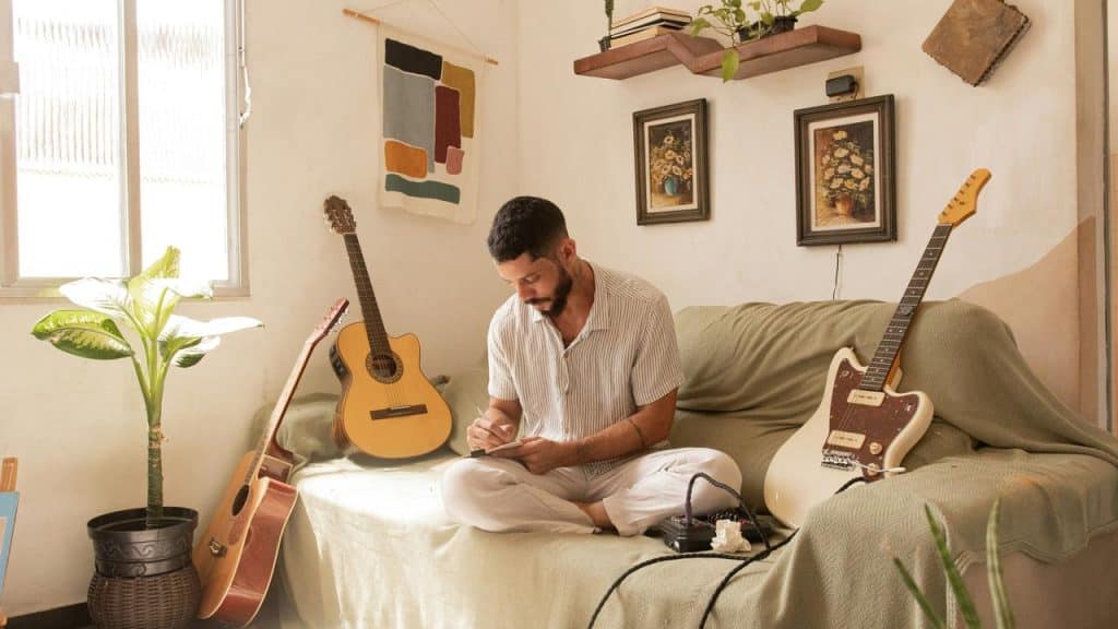 A man writing in a notebook on a sofa surrounded by guitars.