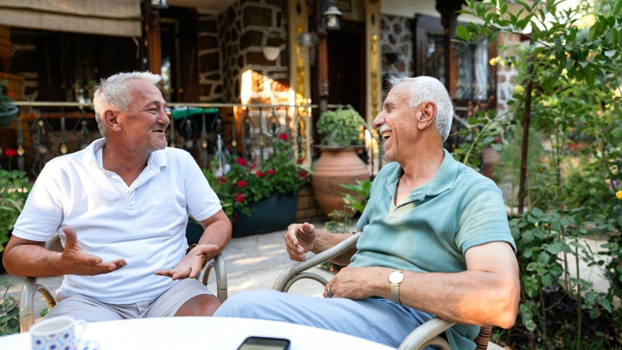 Two older men, one in a white polo and one in a teal polo, laugh together outdoors.