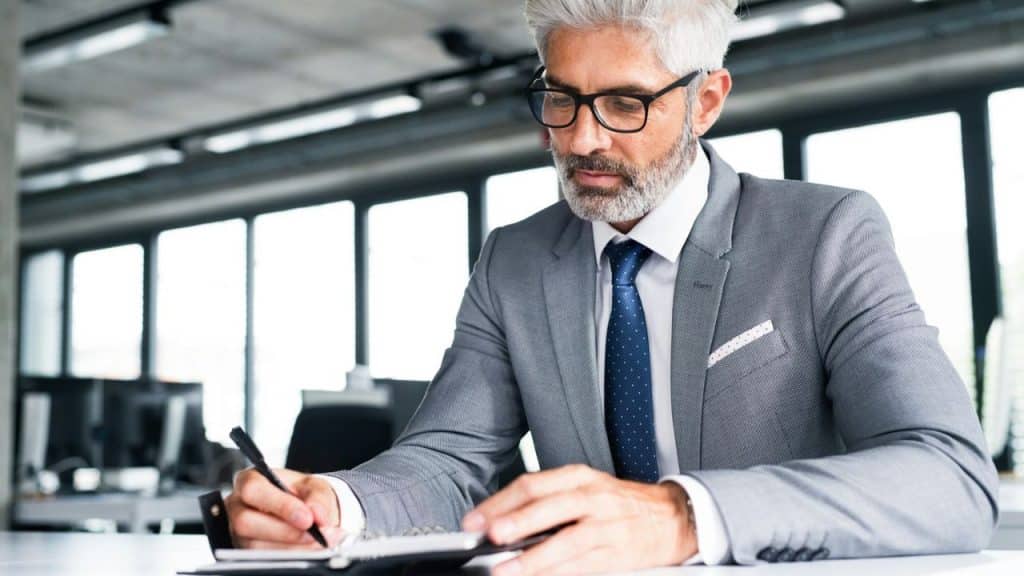A grey-haired man with a beard and glasses, wearing a suit, writes in a notebook.