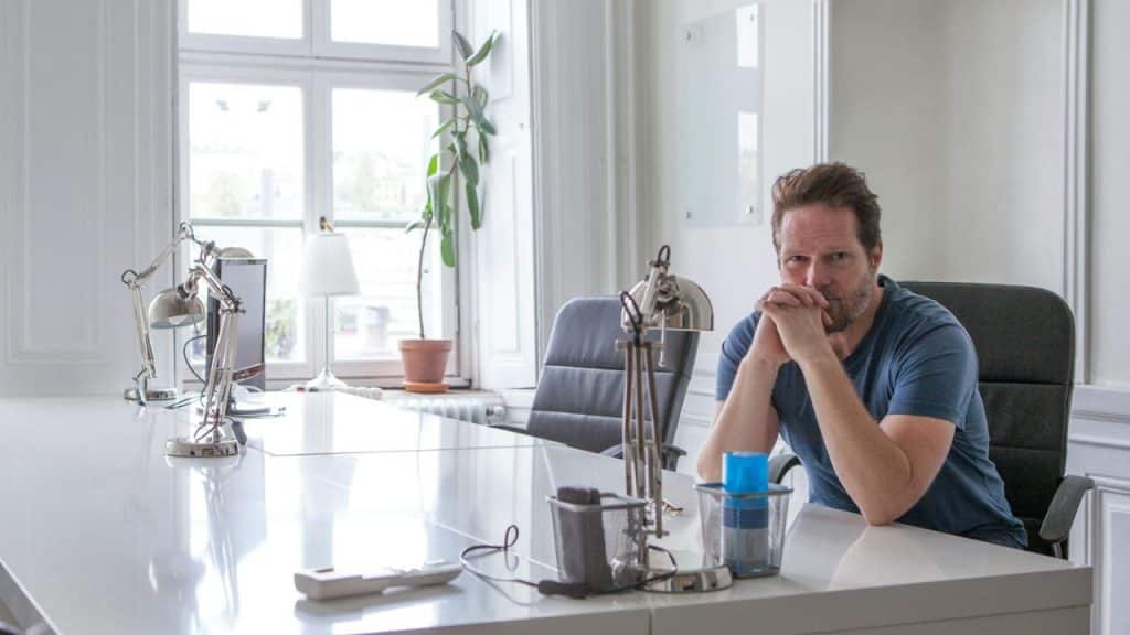 A man with his hands clasped under his chin sits at a desk in a bright room.