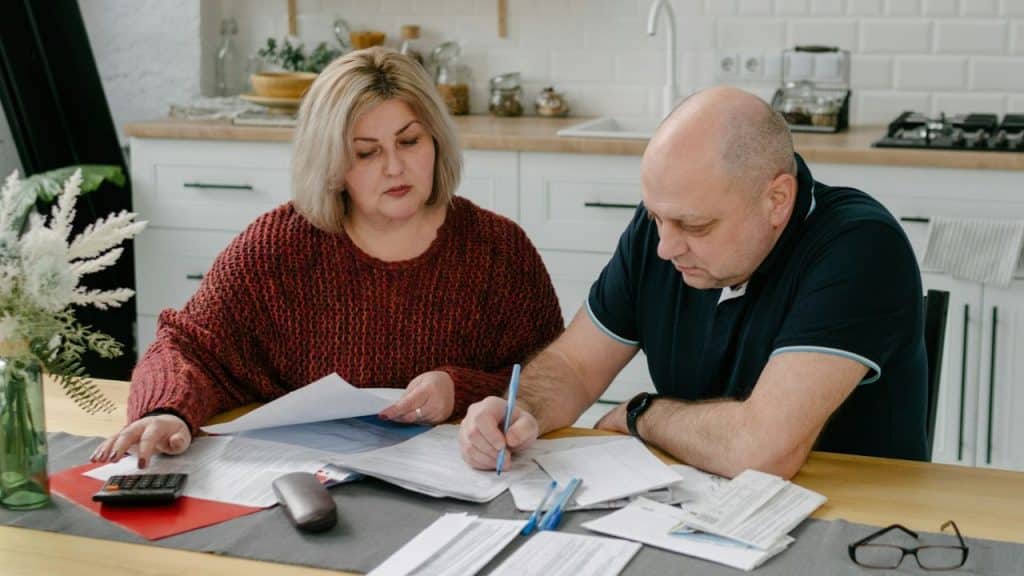 A serious middle-aged couple reviews documents and finances at a kitchen table.