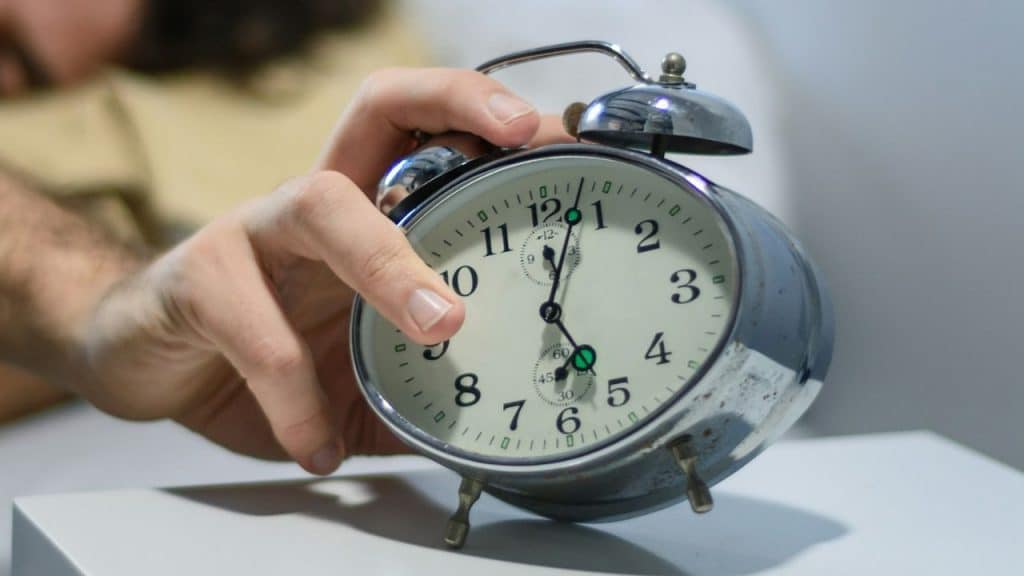 A person's hand reaches for an old-fashioned alarm clock on a bedside table.