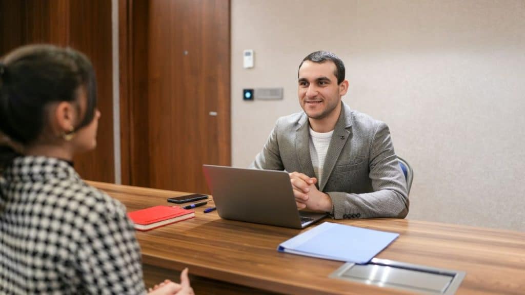 A smiling man in a grey jacket sits at a desk with a laptop, facing a person with their back to the camera.