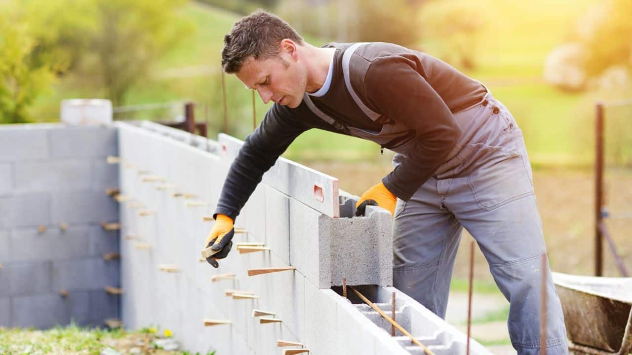A man in work clothes and gloves lays concrete blocks outdoors, building a wall.