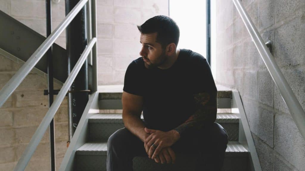 A man with a beard sitting on stairs in a dimly lit stairwell, looking to the side.