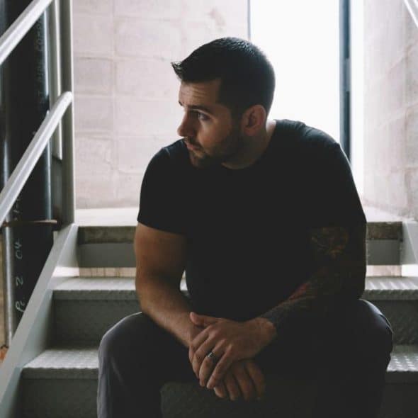 A man with a beard sitting on stairs in a dimly lit stairwell, looking to the side.