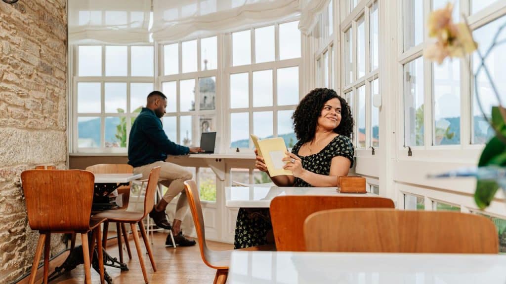 A woman reading a book by a window table as a man works on a laptop in a bright café.