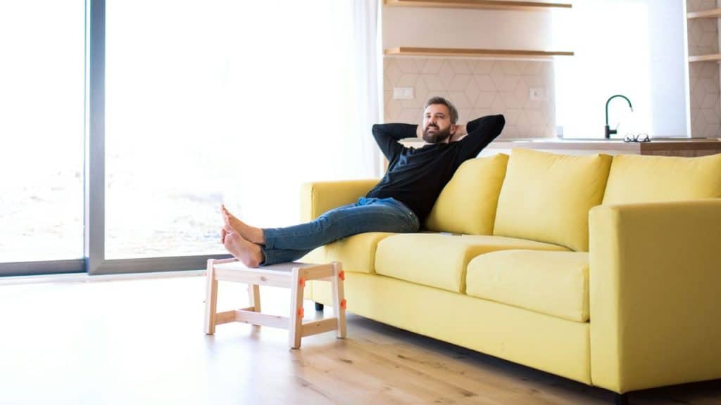 A relaxed man reclines on a yellow sofa with his feet up, gazing out a window.