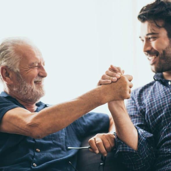 An older man and a younger man smiling as they clasp hands on a sofa.