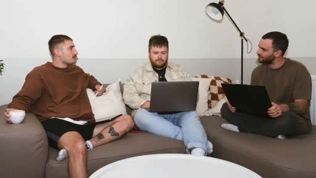 Three men on a couch, two with laptops, one holding a tablet and coffee.