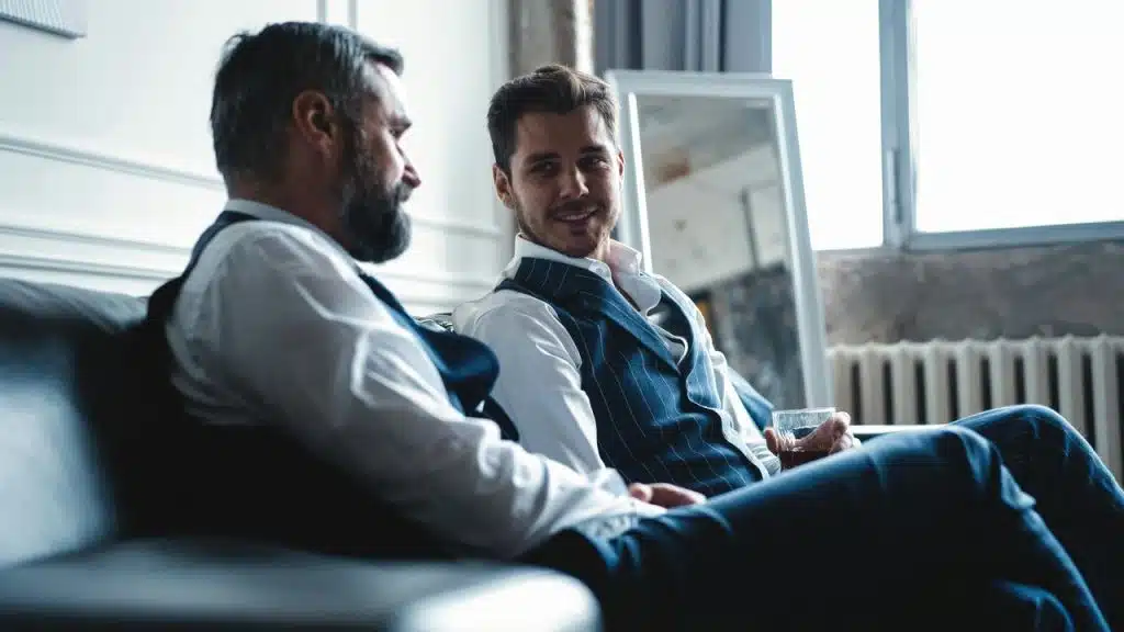 A young man and an older man in vests talking on a sofa.