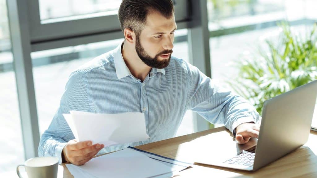 A bearded man holding papers and working on a laptop at a sunlit desk.
