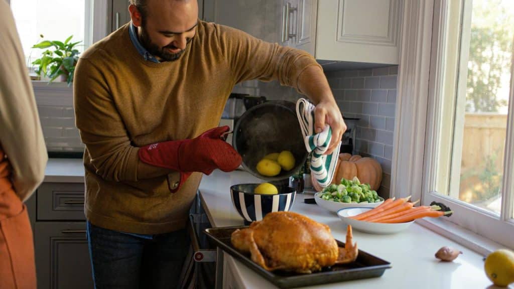A man pouring cooked potatoes into a bowl beside a roasted turkey.