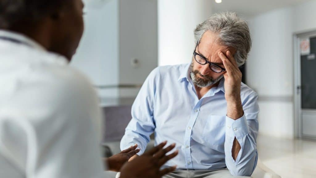 A man with a beard and glasses sits with his hand on his forehead, talking to a person in a white coat.