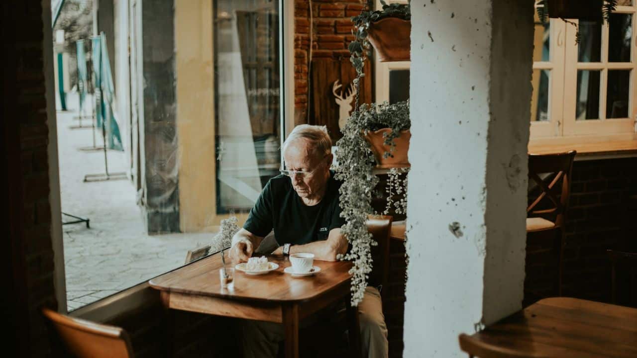 A man with white hair and glasses sits alone at a wooden table in a cafe, looking down at his food.