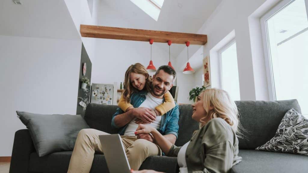 A mother showing her laptop to her husband while their daughter hugs him on the couch.