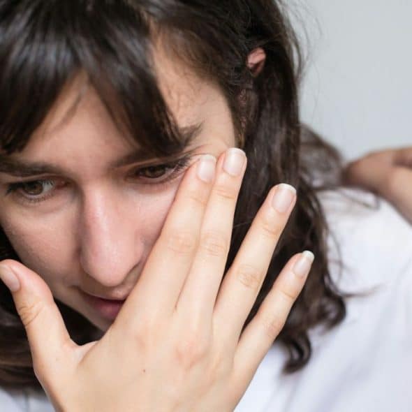 A distressed woman covers part of her face with her hand, while another hand rests on her shoulder.