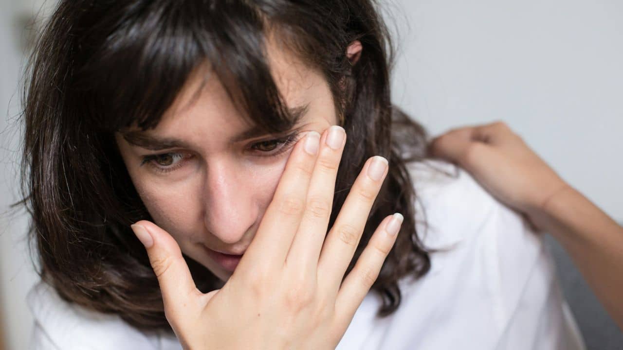 A distressed woman covers part of her face with her hand, while another hand rests on her shoulder.