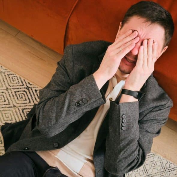 A man in a gray jacket lies on a patterned rug, covering his eyes with his hands.