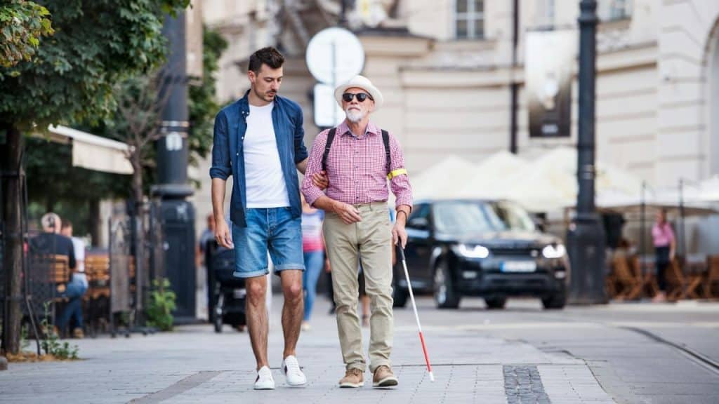 A young man walks alongside an older man with a white cane and hat, guiding him.