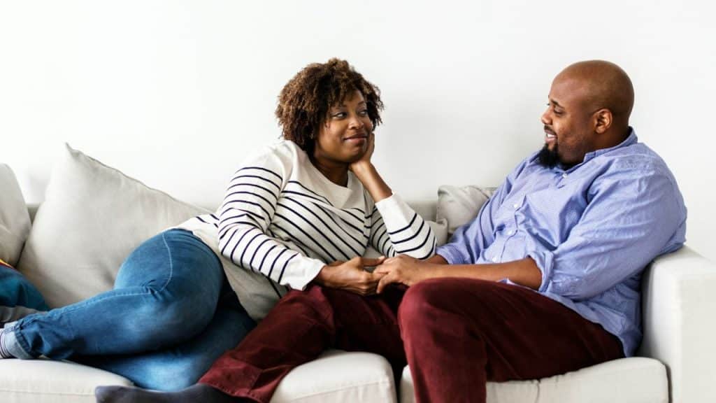 A couple smiles at each other while relaxing on a white sofa.