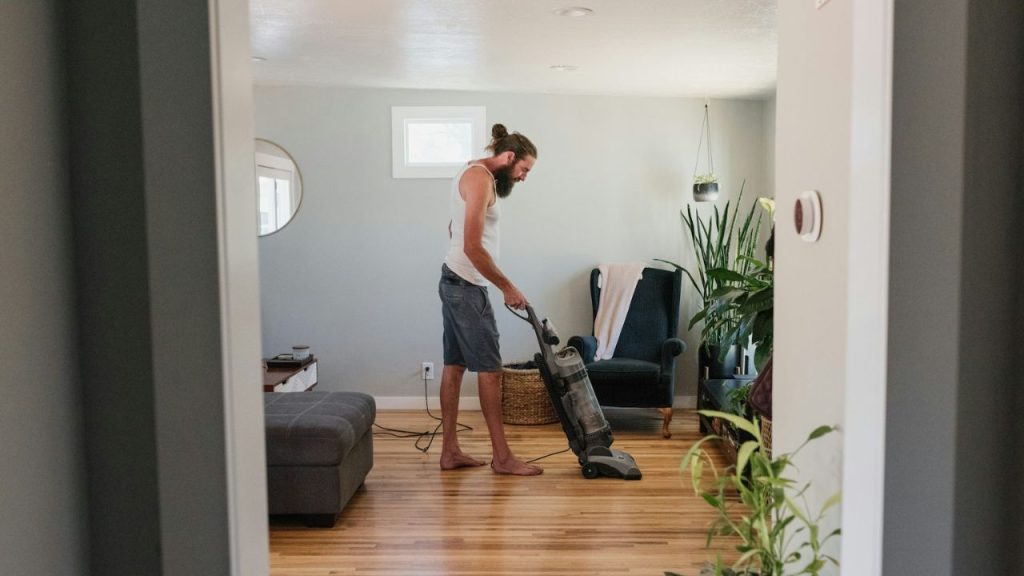 A man with long hair in a bun vacuums a hardwood floor in a living room.