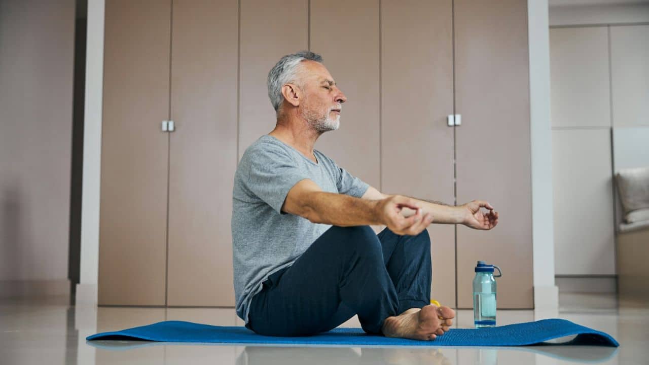 A senior man meditates on a blue yoga mat with a water bottle nearby.