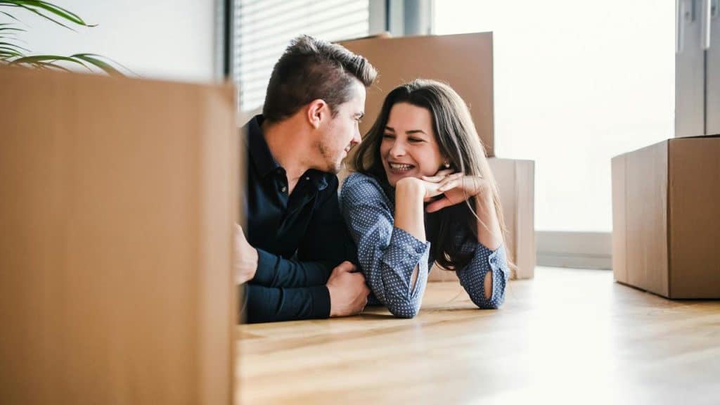 A couple smiling on the floor amid cardboard moving boxes.