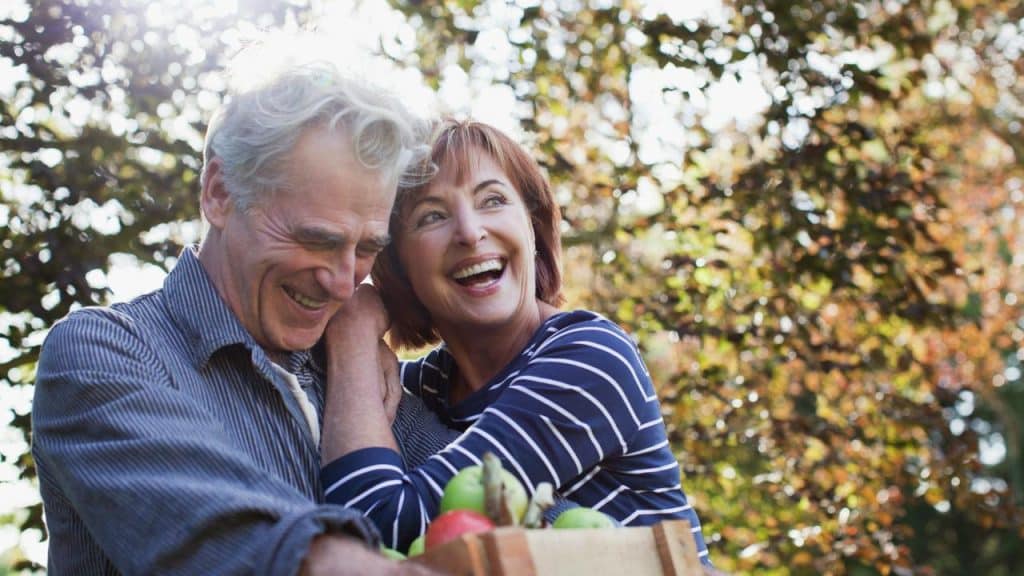 A laughing older couple holding a crate of apples outdoors.