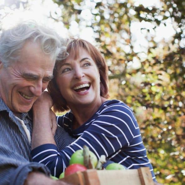 A laughing older couple holding a crate of apples outdoors.