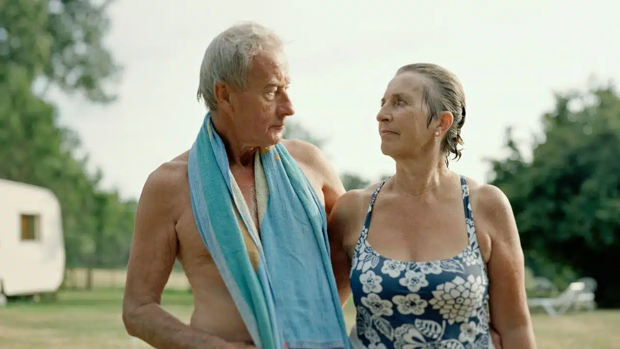A senior couple in swimwear, with a towel draped over the man, stands outdoors looking at each other.