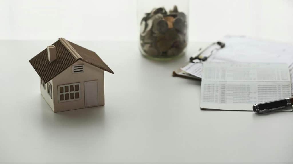 A model house beside paperwork, a pen, and a jar of coins on a white desk.