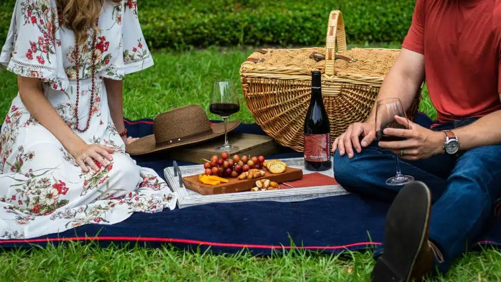 A picnic with wine, cheese, and a wicker basket beside two people on a blanket.