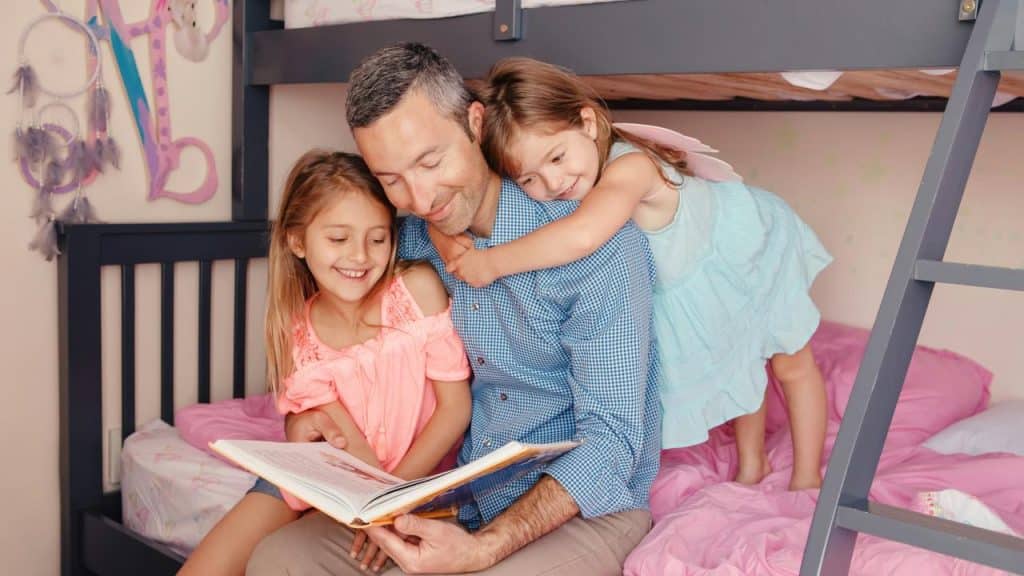 A father reading a book to his two daughters on a bunk bed.