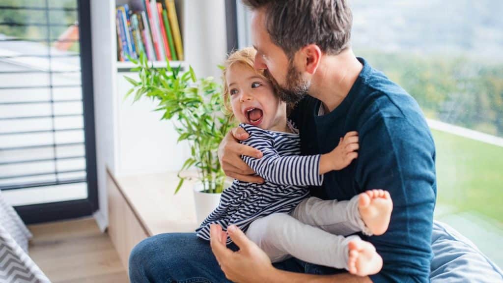 A father playfully hugging his laughing toddler by a window.