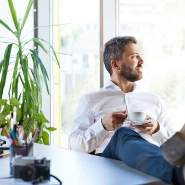 A bearded man in a white shirt and blue jeans, with his feet up on a desk, sips from a cup while looking out a window.